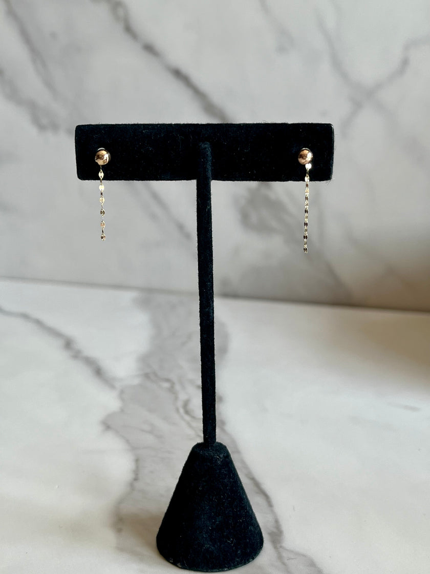 A pair of gold dangle earrings with small round studs and dangling chain links, displayed on a black velvet jewelry stand against a white marble background.